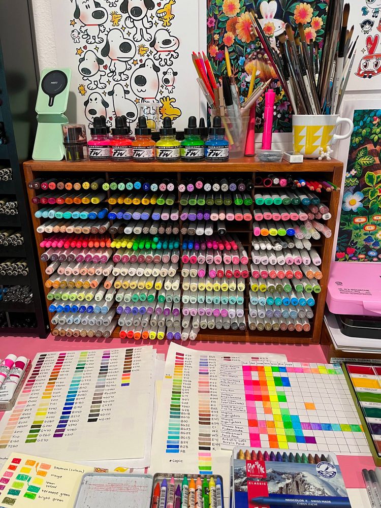 An image of a desk with two large pen displays on it. The display have hundreds of markers. There are swatch cards and other art supplies (paints and pastels) on the table as well 