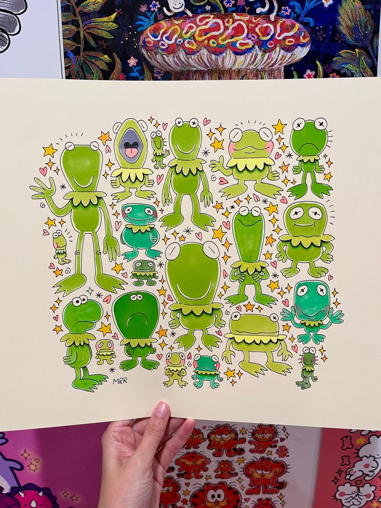 A hand is holding up a paper with an illustration on it! The illustration features 20 Kermit drawings. They’re a little wonky!! They are all different shades of green and yellow green. There are stars and hearts surrounding the Kermits. The drawing is with ink and alcohol makers. 