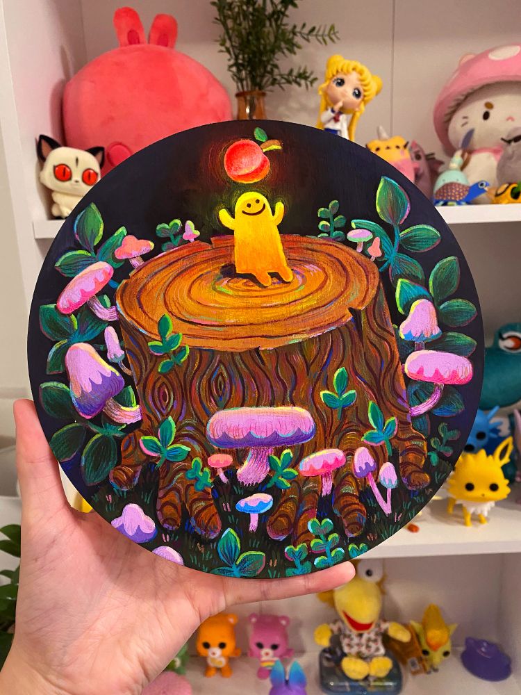 An acrylic painting on a wood round. A little yellow creature is sitting on top of a tree trunk, and there's a floating peach above him. Behind the tree trunk are leaves and mushrooms!