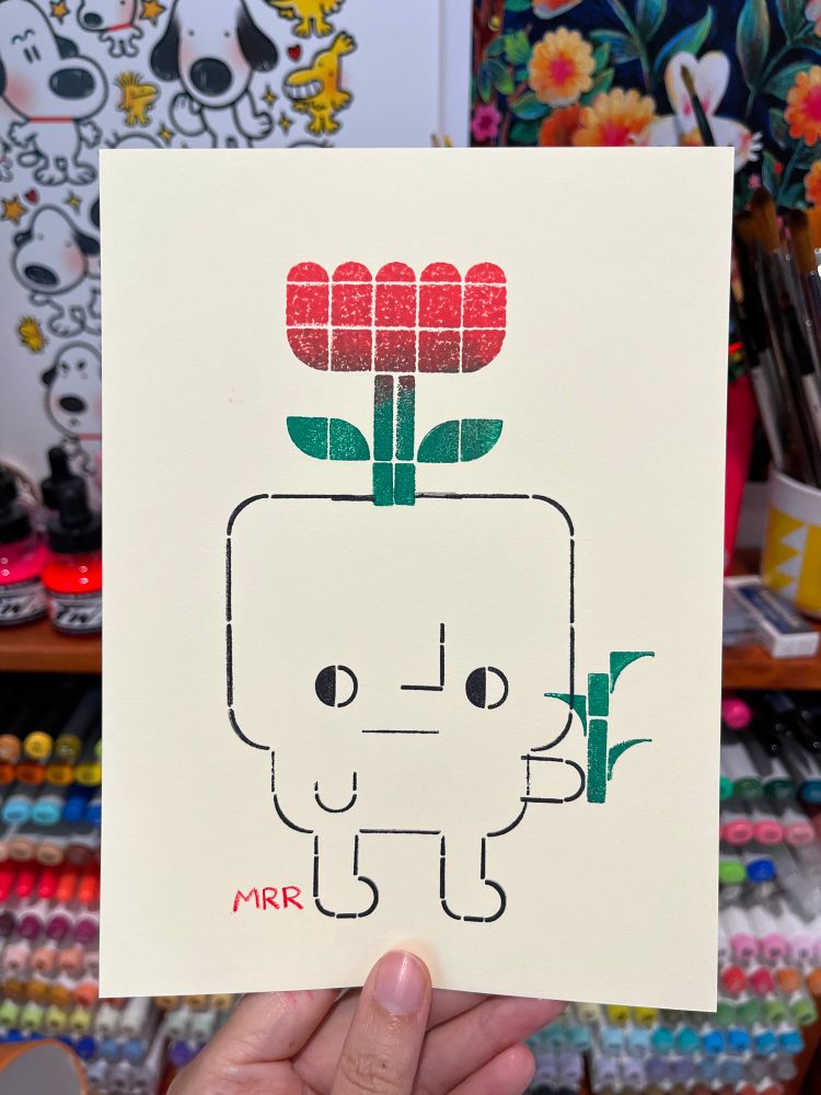 A hand holding up a small card with a block print on it. The print is of a guy holding a plant. The guy also has a flower growing out of his head 