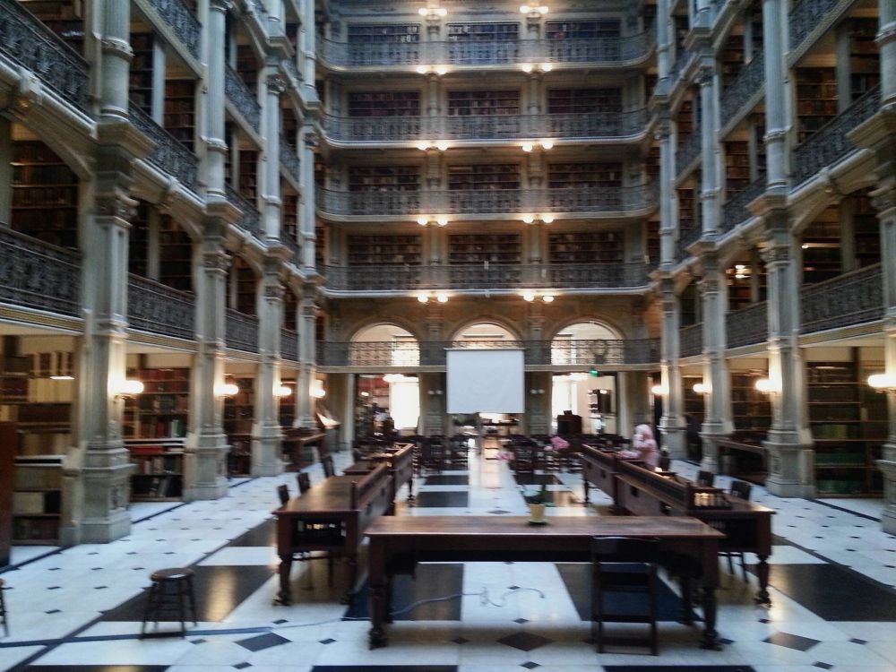 The interior of a large library. Study tables line a marble floor. Pillared open floors ascend four stories.