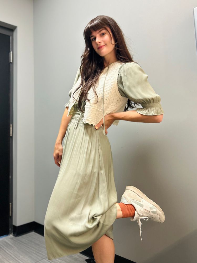 OP showing her outfit with her foot kicked up—sage green dress, cream knit vest, orange socks and Reeboks.