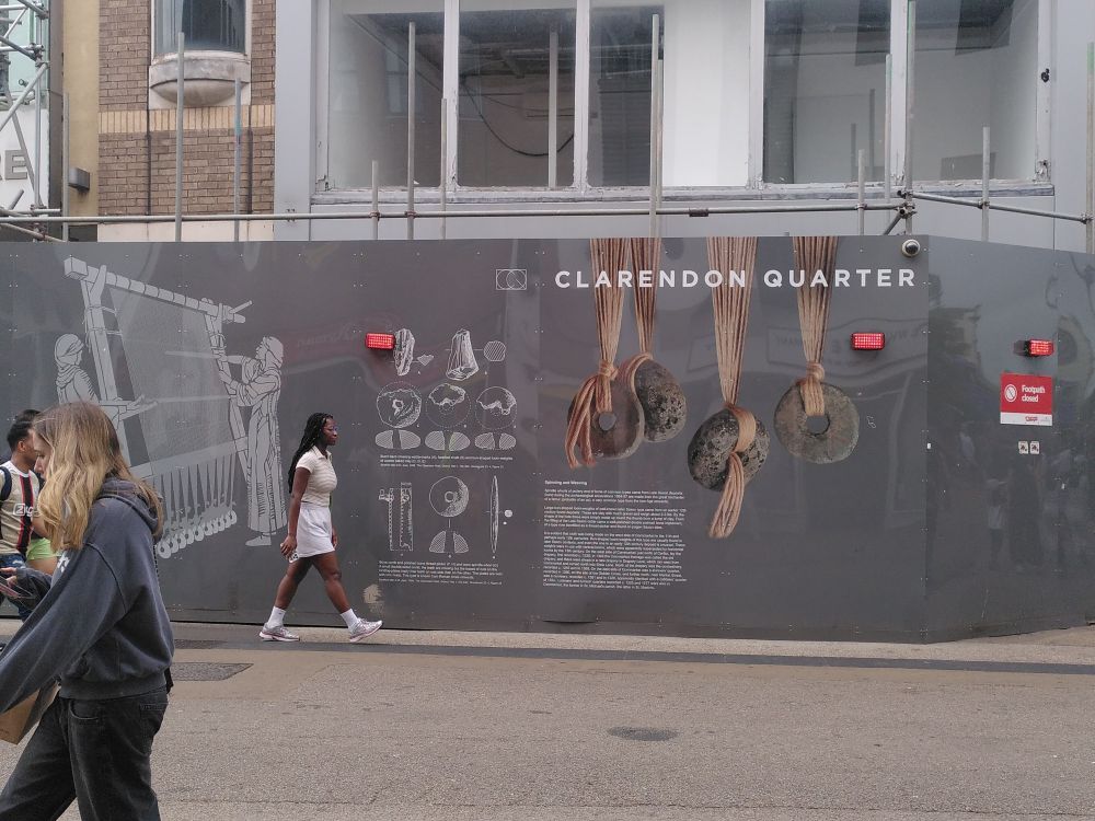 Photo of a street with people walking by a large hoarding title Clarendon Quarter. Below are some large photos of loom weights, and line drawings of a loom and the finds, with text.
