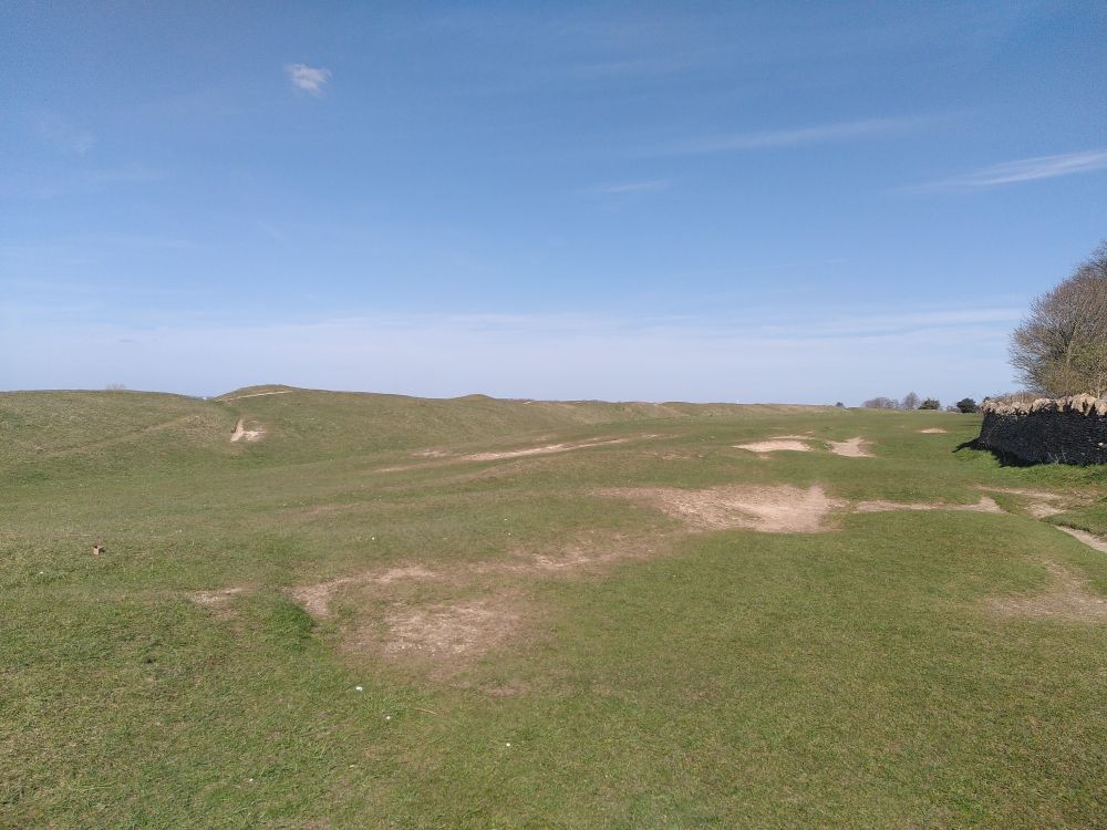 A view across veryand bare patches towards a long bank stretching across the whole picture. A ditch just visible on this side. A ridiculously sunny day.