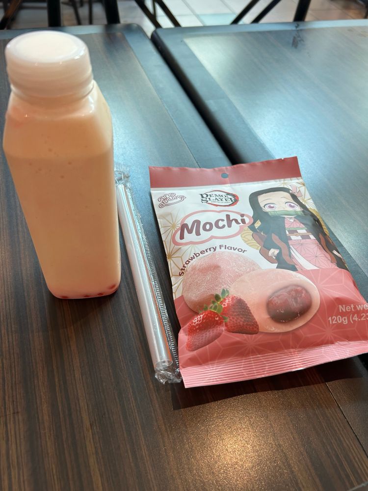 this is a photo of my pale orange-coloured peach smoothie with red strawberry popping boba at the bottom of it, sitting next to a pink bag of strawberry mochi. the mochi bag has an image of nezuko kamado from demon slayer on it, as well as the demon slayer logo and images of the strawberry mochi.