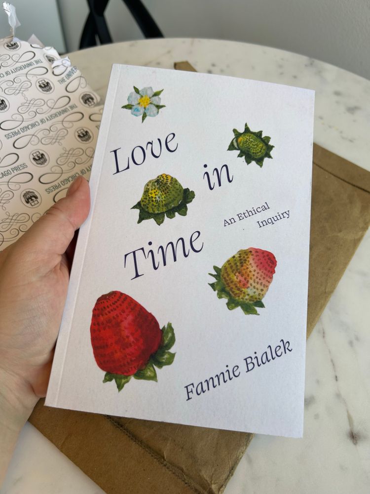Hand holding copy of book called Love in Time by Fannie Bialek. Cover is off white with five hand painted strawberries at different stages of ripeness, from flower to fully red and ready to eat. 
