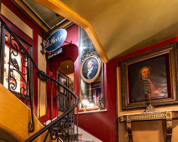 scala interna di caffè antico parigino, con soffitti a volta ocra, pareti rosso scuro e quadri appesi; si vede anche l'ingresso a una saletta.
Foto da internet