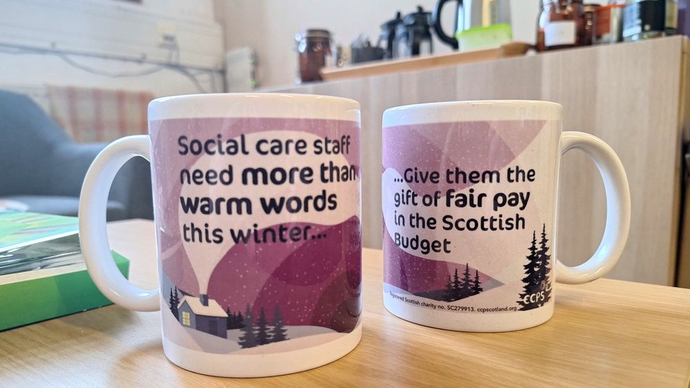 Two mugs sitting on a table. The mugs have a berry coloured snow scene on them. There is wording on the mugs which reads 'Social care staff need more than warm words this winter ... Give them the gift of fair pay in the Scottish Budget.'