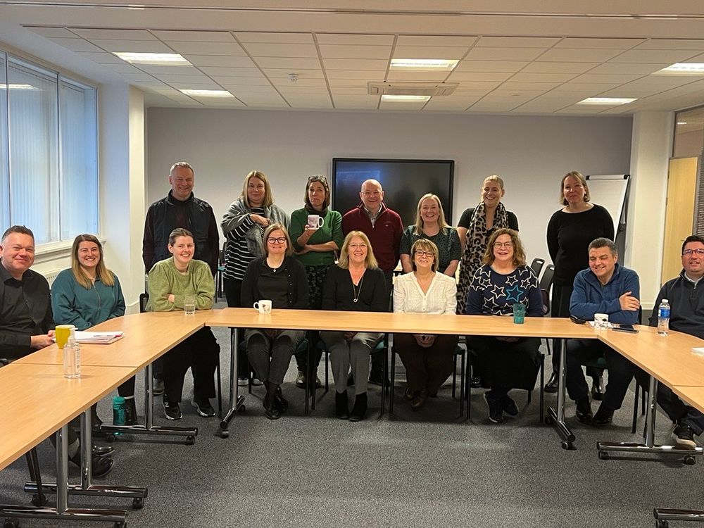 A group of people around a boardroom style table. Some are standing and others are sitting. They are all looking towards the camera and smiling.
