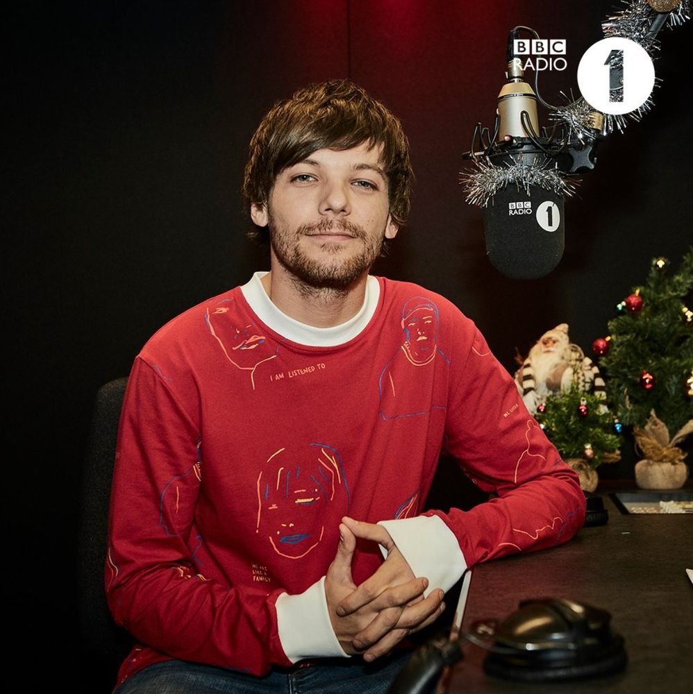 Louis Tomlinson, seated, supposedly in a BBC studio for Christmas. Behind him are Christmassy images and tinsel around a microphone. He wears a red sweater. It's November. 