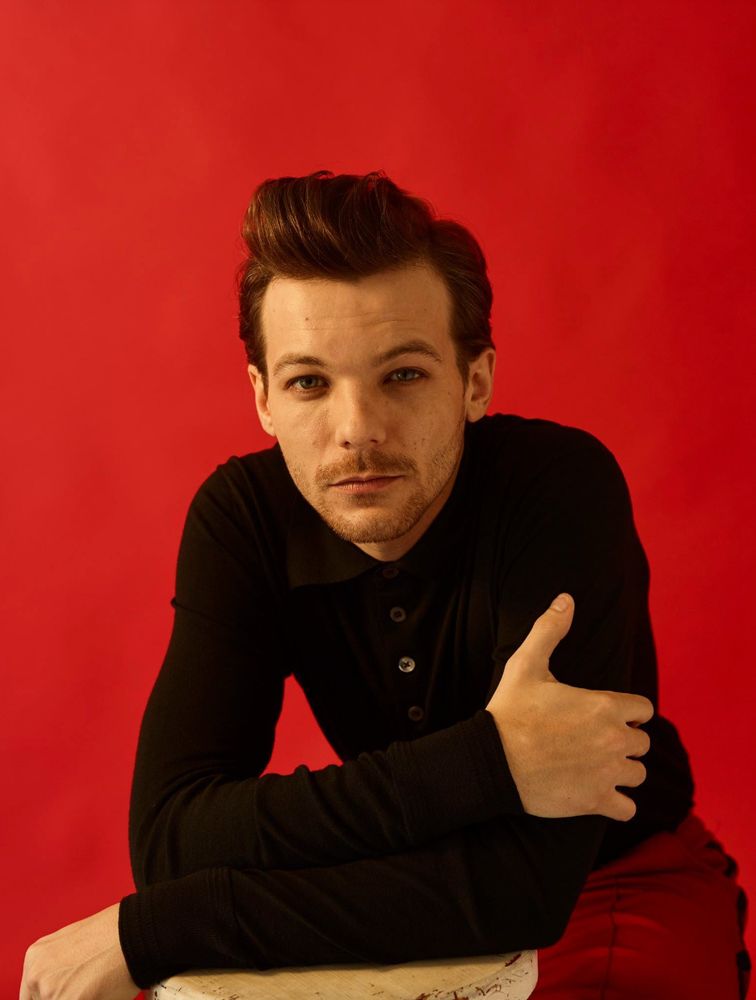Louis Tomlinson in a black shirt, looking directly at the camera. His hair is in a quiff. He is actually squatting on the floor and leaning on a stool, which is just visible in the foregroun