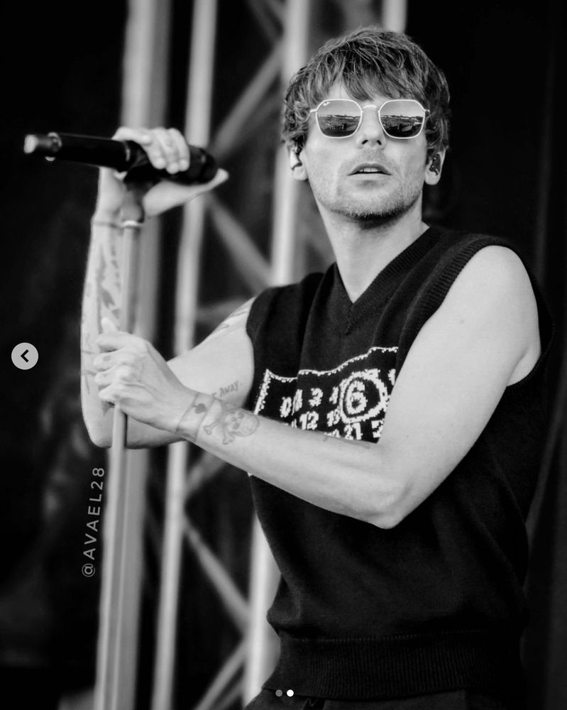 Mono image of Louis Tomlinson on stage wearing a black sweater vest with white writing, and sunglasses. He is clutching the mic and mic stand and looking into the distance.