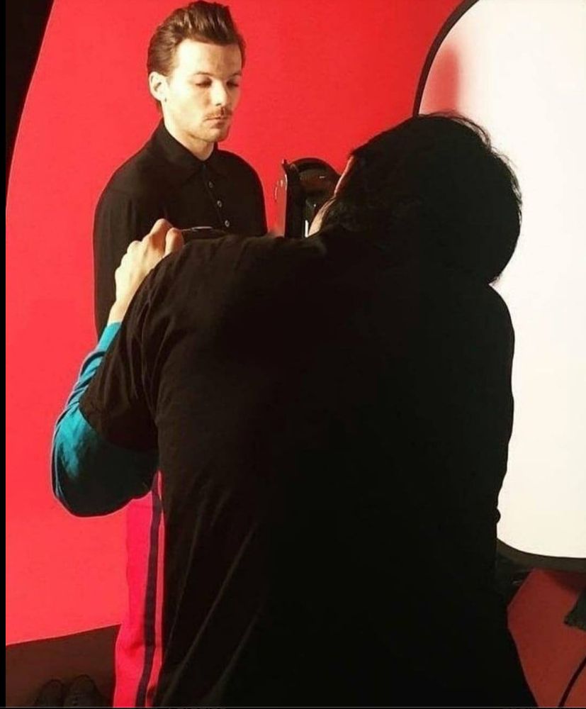 Louis Tomlinson being photographed by a man in black. He is standing in front of a red backdrop. 