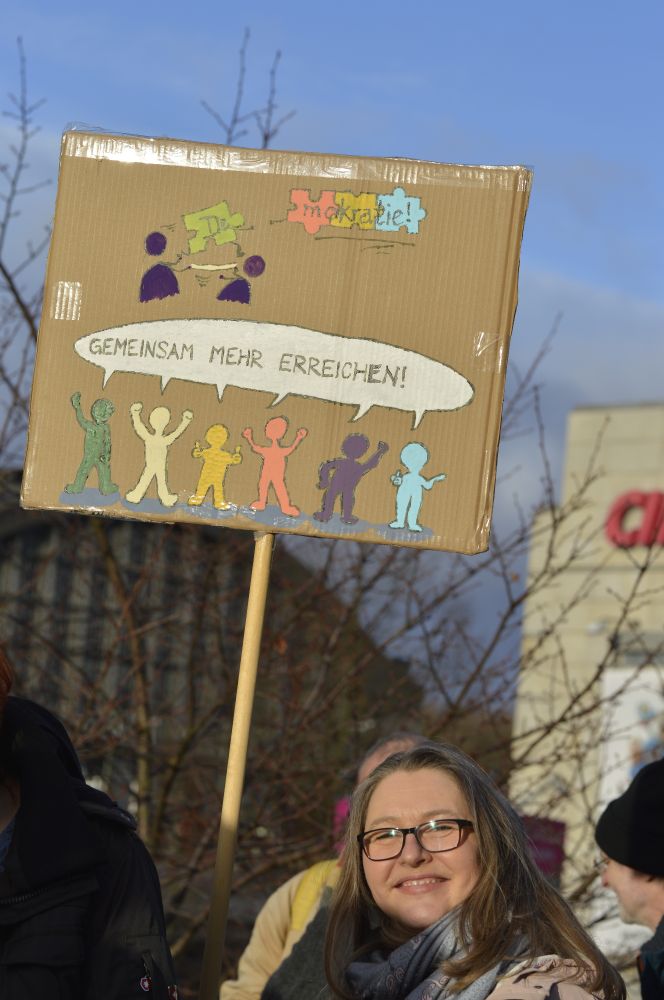 Eine Frau auf der PRÜF-Demo. Sie hält ein Schild hoch. Darauf zu sehen sind Puzzleteile, die gemeinsam das Wort "Demokratie" ausschreiben und Menschen in bunten Farben, die eine gemeinsame Sprechblase über sich haben. In der Sprechblase steht "Gemeinsam mehr erreichen!"