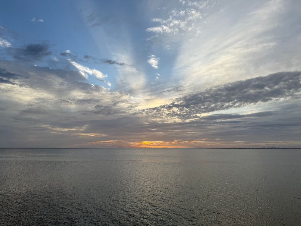 Sunset obscured by low clouds over a calm Mobile Bay, with crepuscular rays shining above the clouds