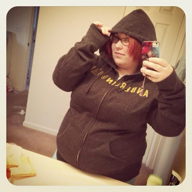 Mirror selfie of a woman in a dark grey zip up hoodie, with “Anberlin Vital” across the chest in yellow. The hoodie looks like it fits snuggly, and her abdomen is hanging over the bathroom counter slightly