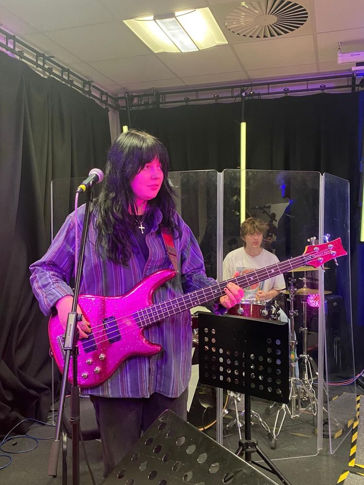 A black-haired girl (my daughter) playing electric guitar, bathed in bright pink light, in a recording space. There is a guy playing drums behind a screen in the background. 