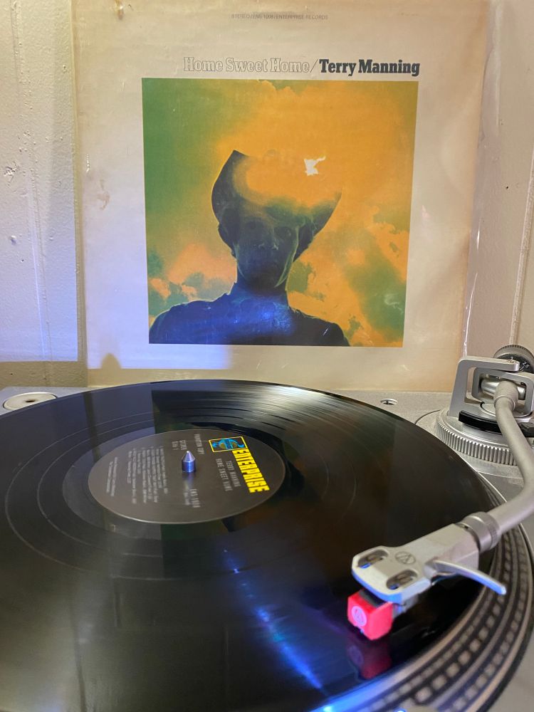 cover of Home Sweet Home by Terry Manning, 1969. A man wearing a hat stands in front of the sun, the sky is yellow and green. Black label Enterprise records label on the record on the turntable 