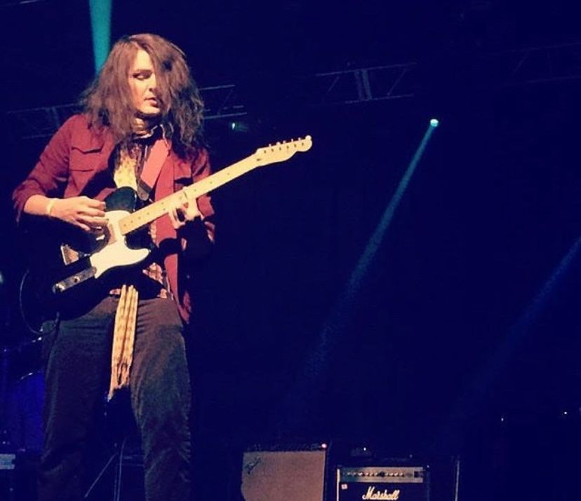 a long haired guy playing a fender telecaster wearing a red jacket, yellow scarf and brown pants with mostly black space, a couple small beams of light and two guitar amplifiers to the right 