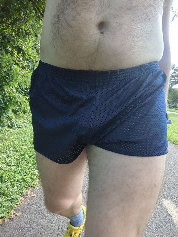 A close up of dude wearing a pair of navy blue running shorts and yellow running shoes walking on a bike trail 
