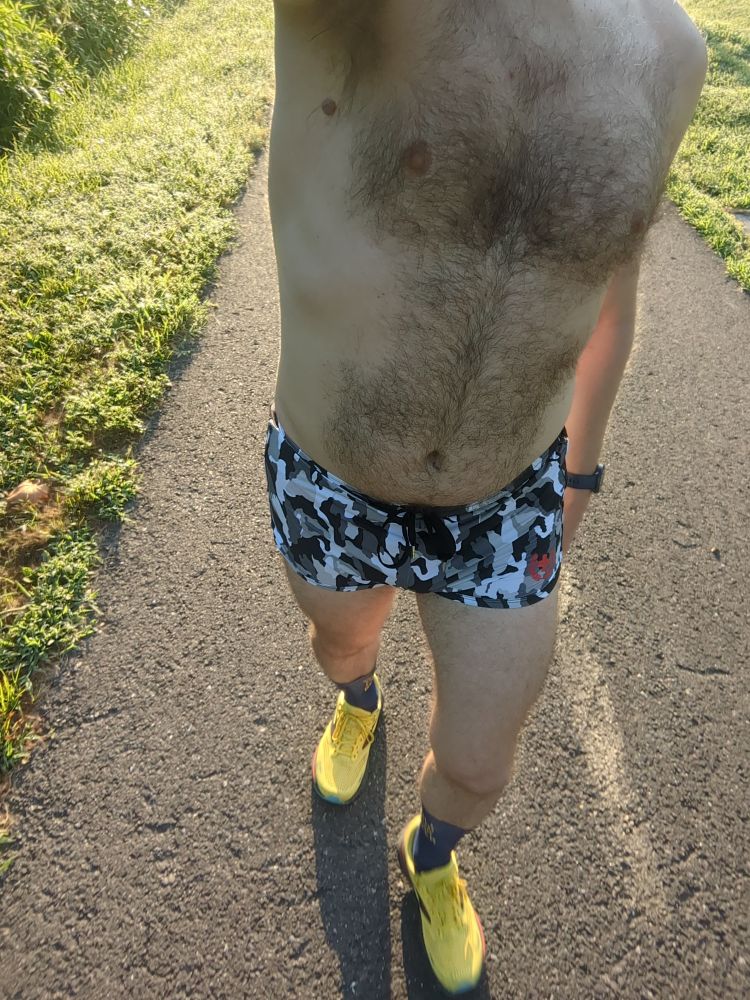 A dude wearing camouflage print running shorts and yellow running shoes walking on a bike trail 