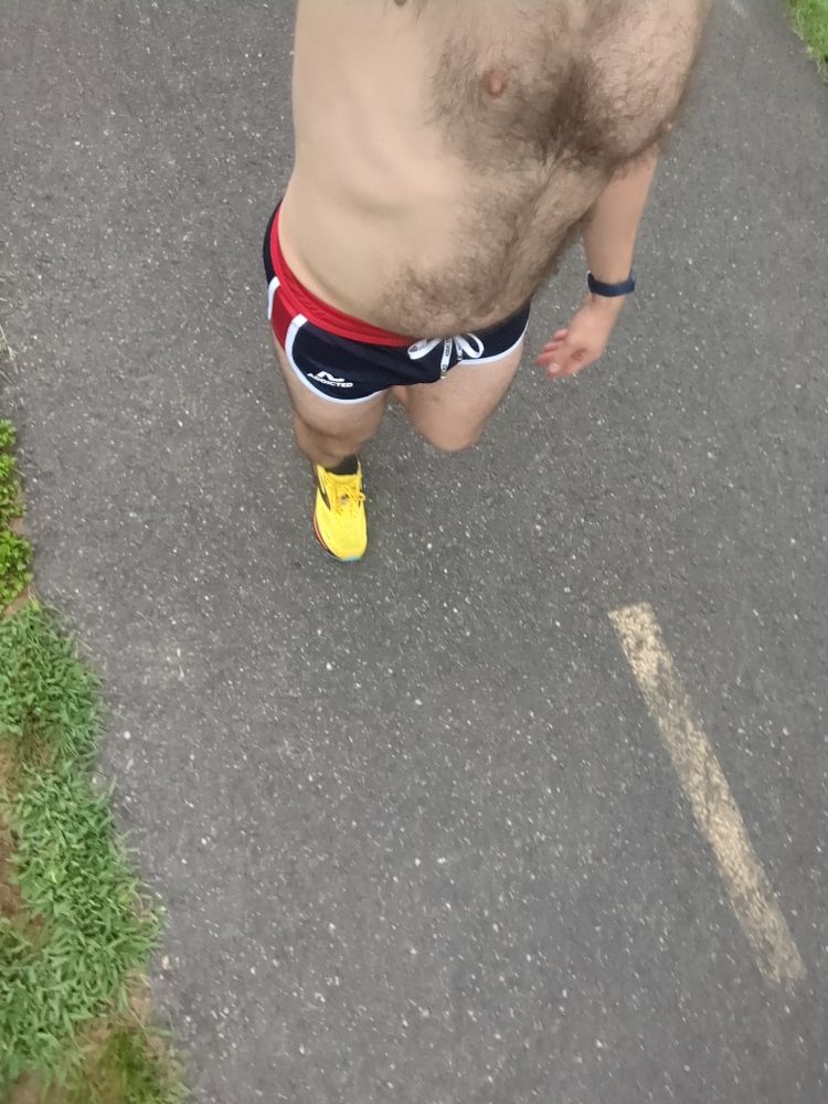 A dude wearing a pair of navy blue running shorts with white and red trim and yellow running shoes walking on a bike trail 