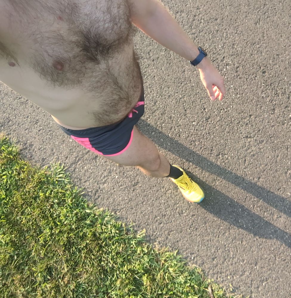 A dude wearing a pair of grey running shorts with neon pink trim and yellow running shoes walking on a bike trail 