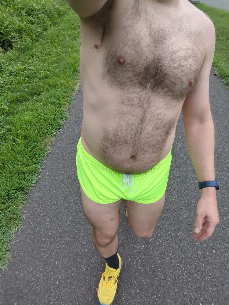 A dude wearing a pair of neon yellow running shorts and yellow running shoes walking on a bike trail 