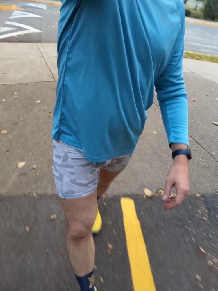 A dude wearing a pair of grey running half tights, blue long sleeve shirt and yellow running shoes walking on a bike trail 