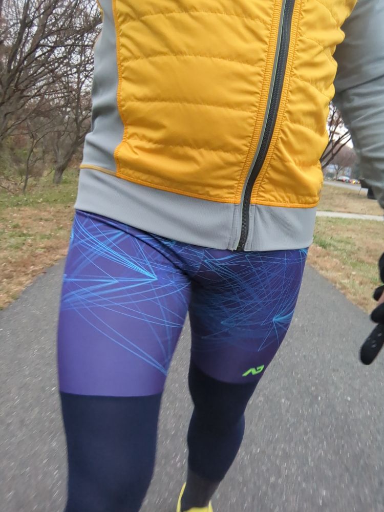 A dude wearing a pair of purple running tights and a yellow running jacket walking on a bike trail 