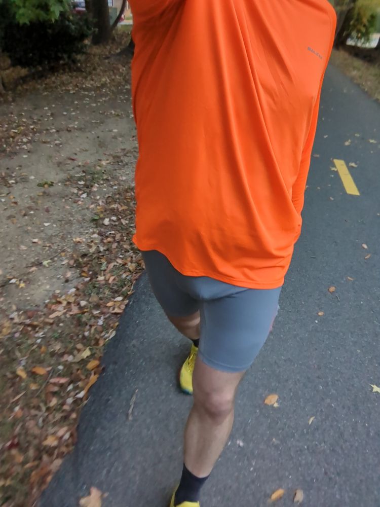 A dude wearing a pair of grey half tights, orange long sleeve shirt and yellow running shoes walking on a bike trail 