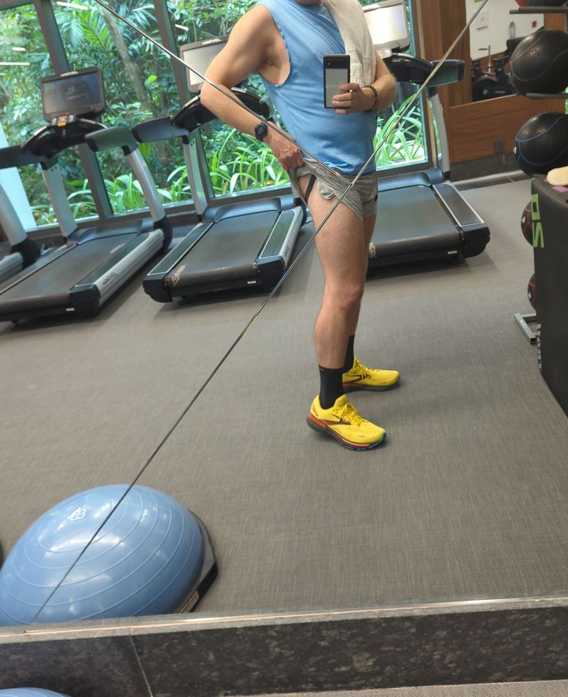 A dude wearing a pair of grey gym shorts, light blue muscle shirt and yellow running shoes taking a selfie in the mirror of a hotel gym while pulling up the hem of one short leg to reveal the strap of a jockstrap 