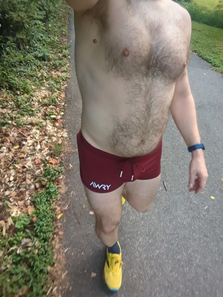 A dude wearing a pair of maroon running shorts and yellow running shoes walking on a bike trail 