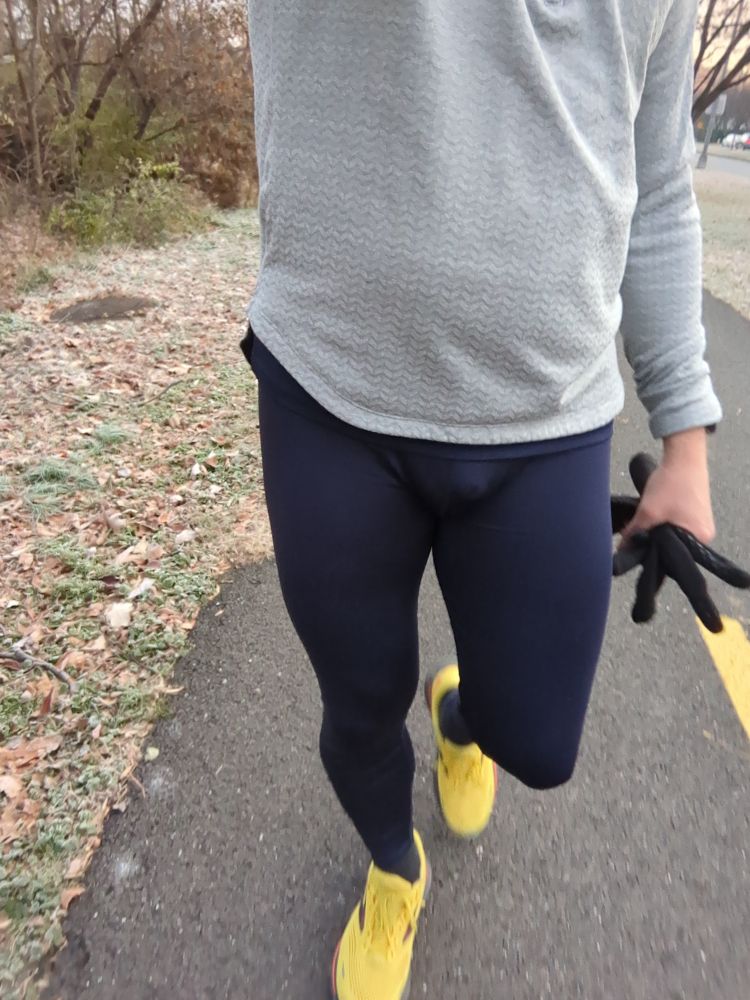 A dude wearing a pair of navy blue running tights, grey running jacket and yellow running shoes walking on a bike trail 