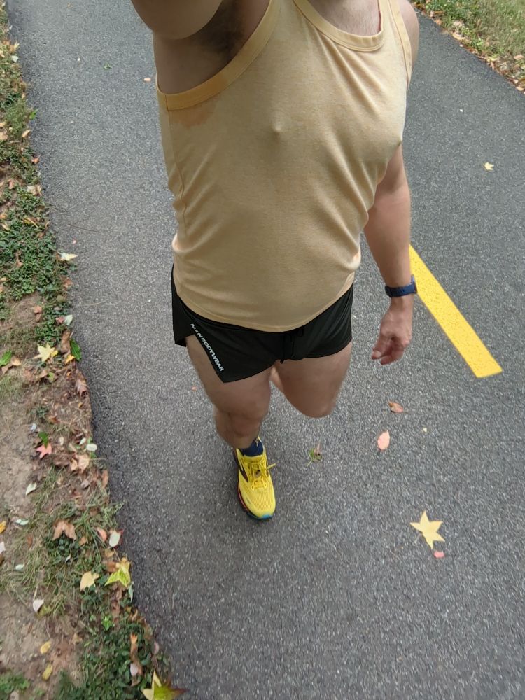 A dude wearing a pair of olive green running shorts, yellow tank top and yellow running shoes walking on a bike trail 