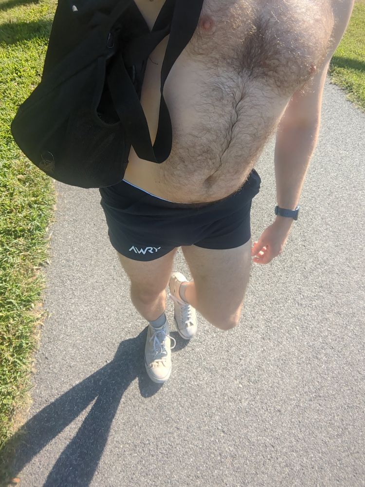 A shirtless dude wearing a pair of navy blue gym shorts and white Chucks walking on a bike trail 