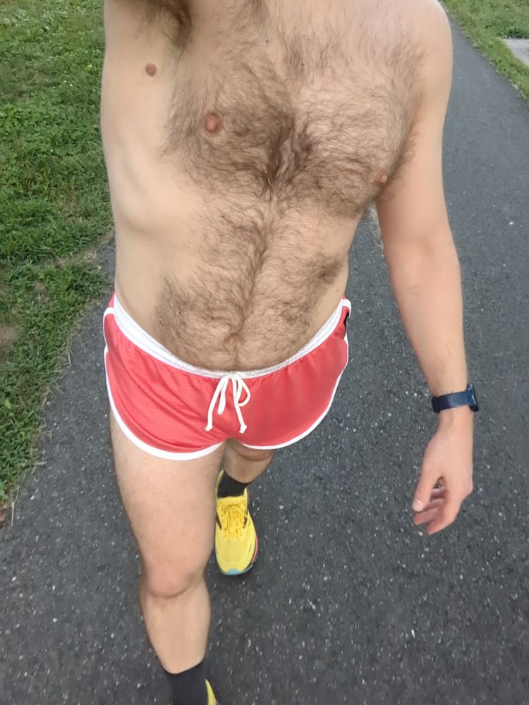 A dude wearing a pair of coral running shorts and yellow running shoes walking on a bike trail 