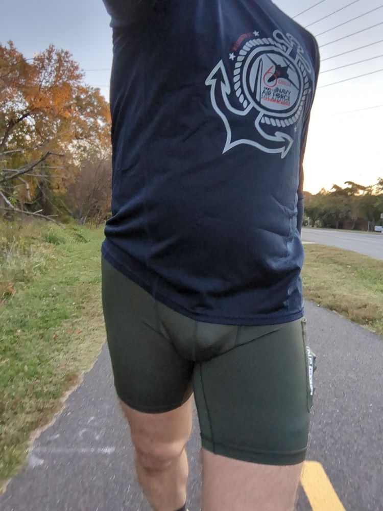 A dude wearing a pair of olive green half tights, navy blue long sleeve shirt and yellow running shoes walking on a bike trail 