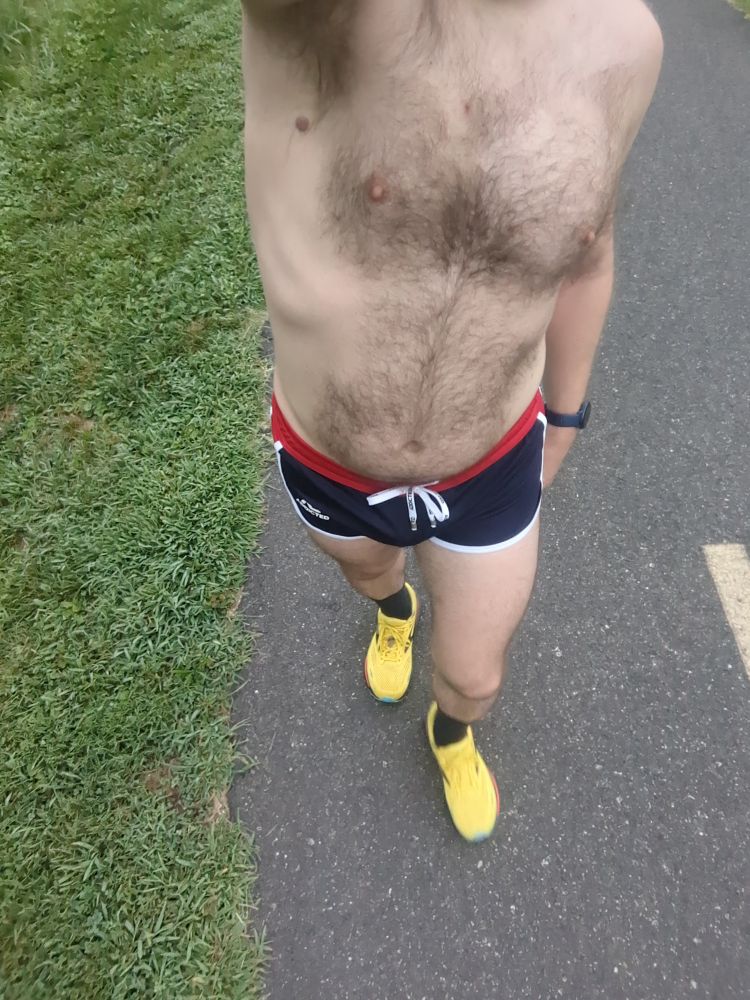 A dude wearing a pair of navy blue running shorts with white and red trim and yellow running shoes walking on a bike trail 