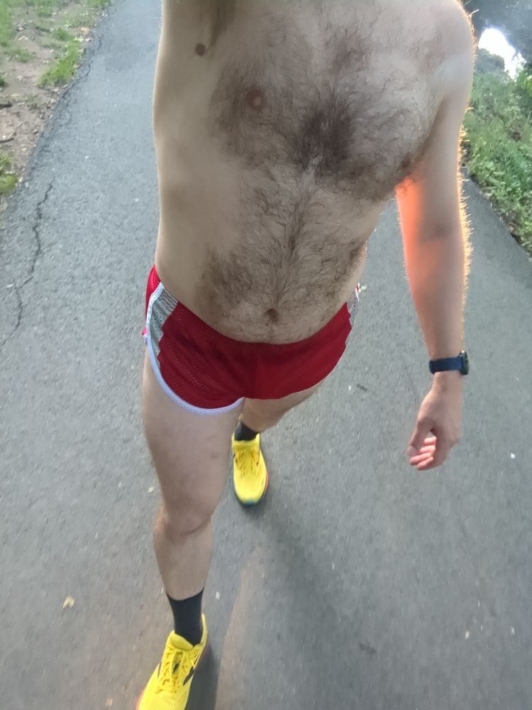 A dude wearing a pair of red running shorts and yellow running shoes walking on a bike trail 