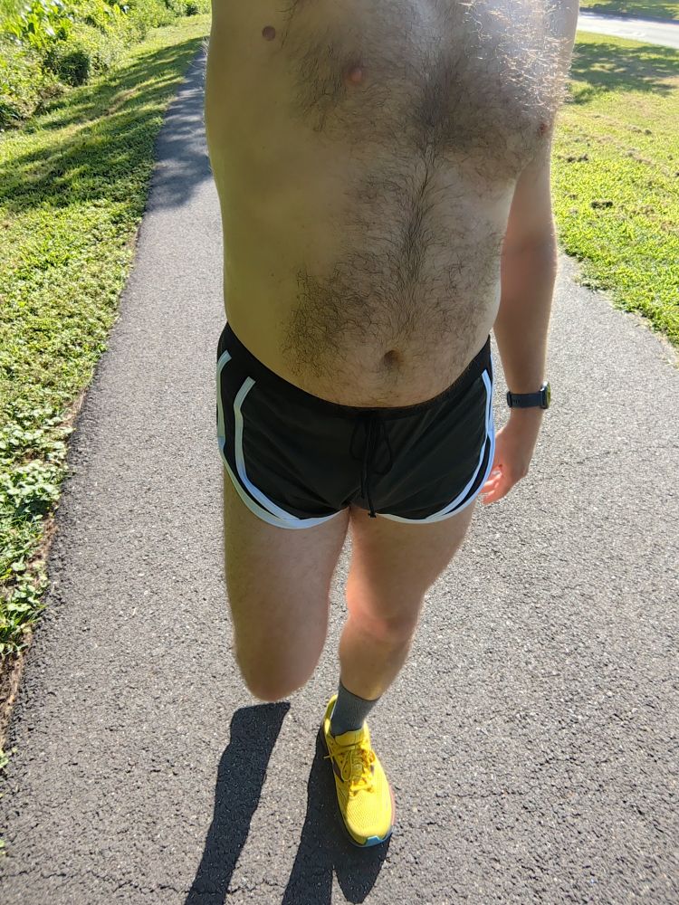 A dude wearing a pair of dark green running shorts with white trim and yellow running shoes walking on a bike trail 