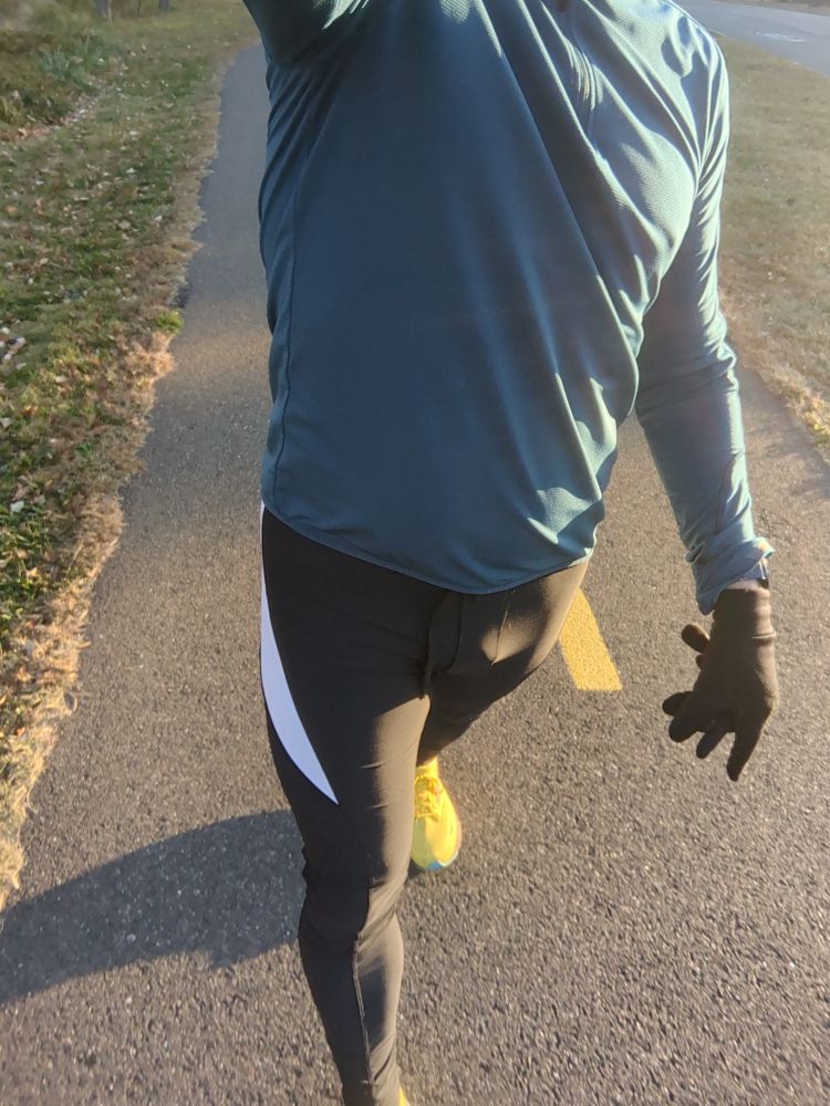 A dude wearing a pair of black running tights, a blue running jacket and yellow running shoes walking on a bike trail 