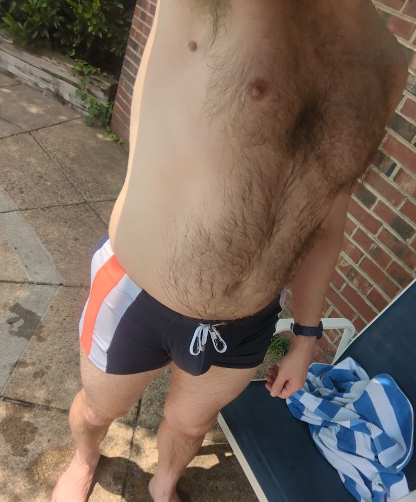 A dude wearing a pair of blue square-cut swim trunks with an orange and white side stripe. He is wet from swimming 