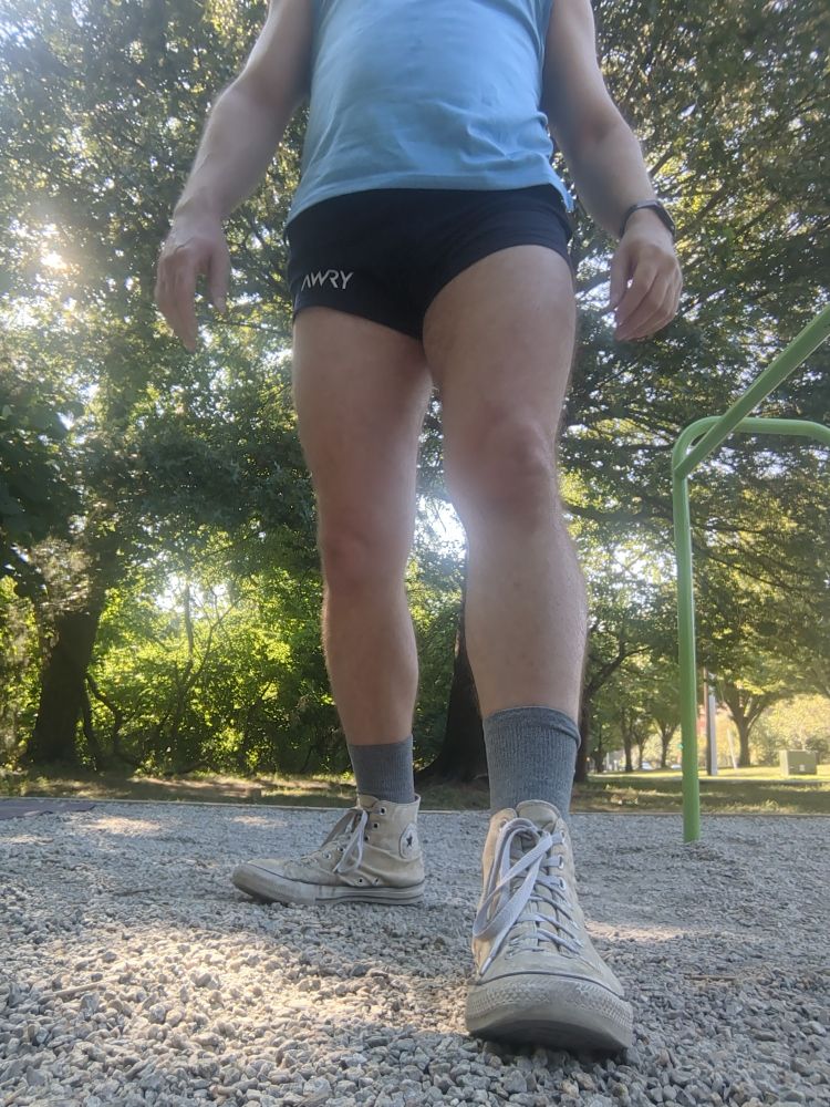 A dude wearing a pair of navy blue gym shorts, light blue muscle shirt and white Chucks standing on a patch of gravel 