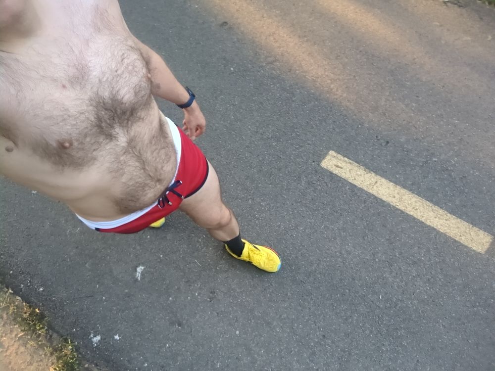 A dude wearing a pair of red and white running shorts and yellow running shoes walking on a bike trail 