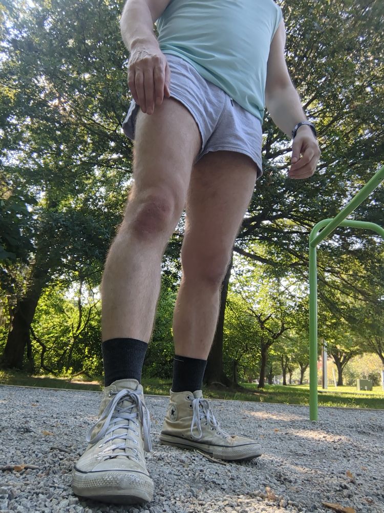A dude wearing a pair of grey gym shorts, light blue muscle shirt and white Chucks standing on a patch of gravel 