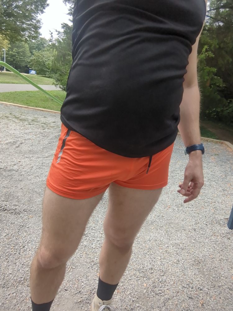 A dude wearing a pair of orange gym shorts and a black muscle shirt standing on a patch of gravel 