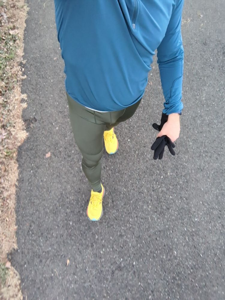 A dude wearing a pair of green running tights, blue running jacket and yellow running shoes walking on a bike trail 