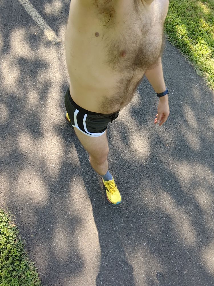 A dude wearing a pair of dark green running shorts with white trim and yellow running shoes walking on a bike trail 