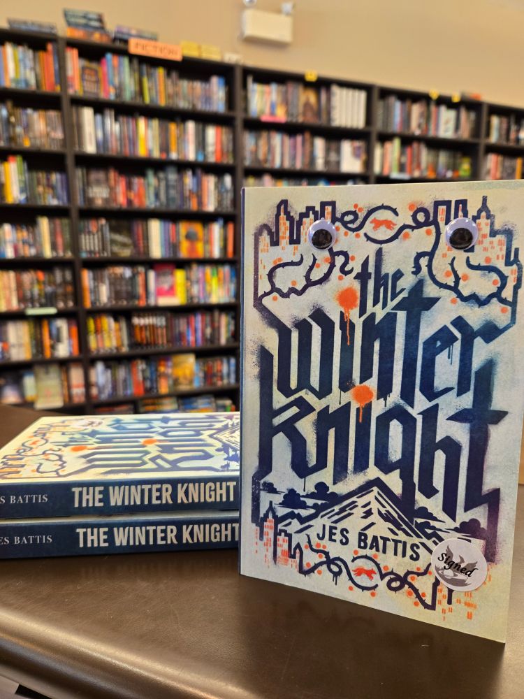 Three copies of the Winter Knight by Jes Battis. One copy is standing up, and has a pair of googly eyes adorning the front cover. 